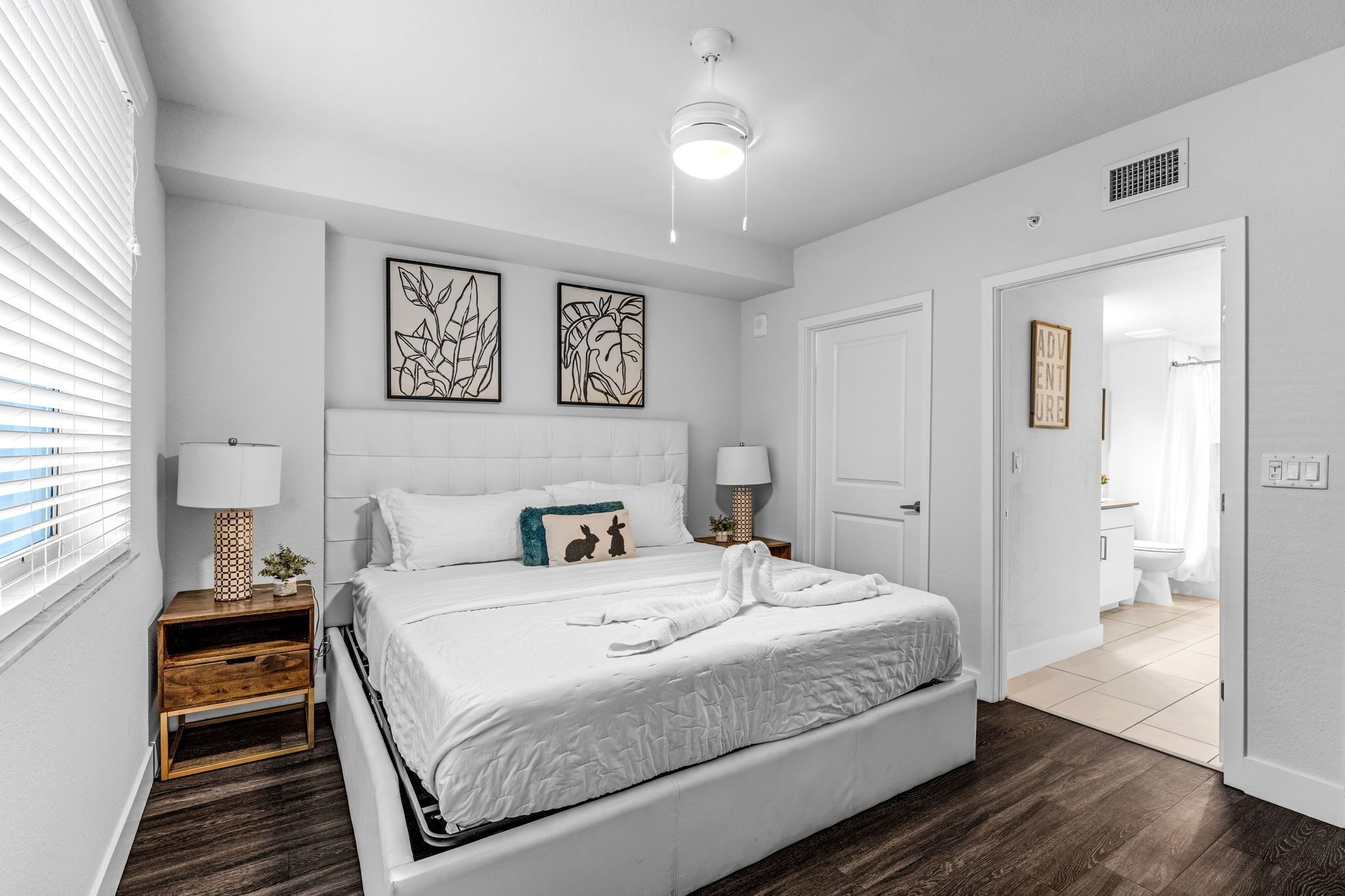 Um quarto com uma cama, duas mesas de cabeceira, um criado-mudo, uma luminária no teto e uma porta que dá para o banheiro.