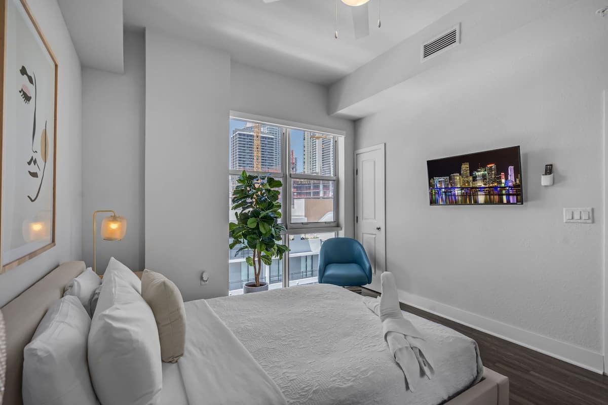 A modern hotel room with a bed, a blue chair, a potted plant, a cityscape view, and a mounted TV.