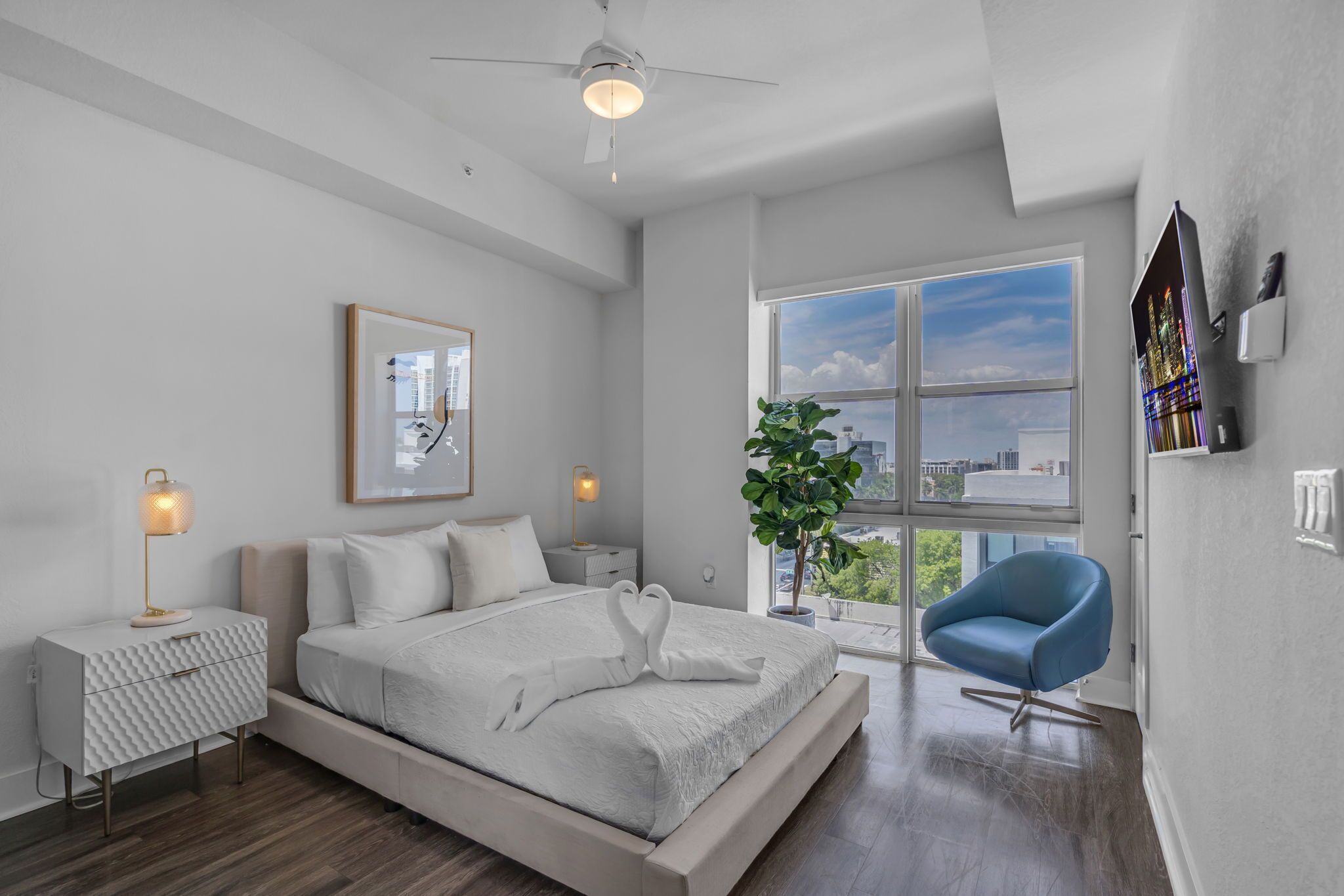 Une chambre moderne avec un grand lit, un ventilateur de plafond, une fenêtre donnant sur la ville, un fauteuil bleu et une plante.