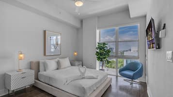 Un dormitorio moderno con una cama grande, un ventilador de techo, una ventana con vistas a la ciudad, una silla azul y una planta.