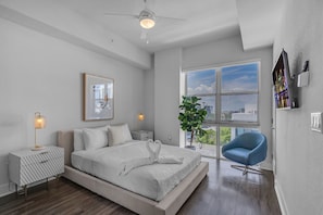 Un dormitorio moderno con una cama grande, un ventilador de techo, una ventana con vistas a la ciudad, una silla azul y una planta.