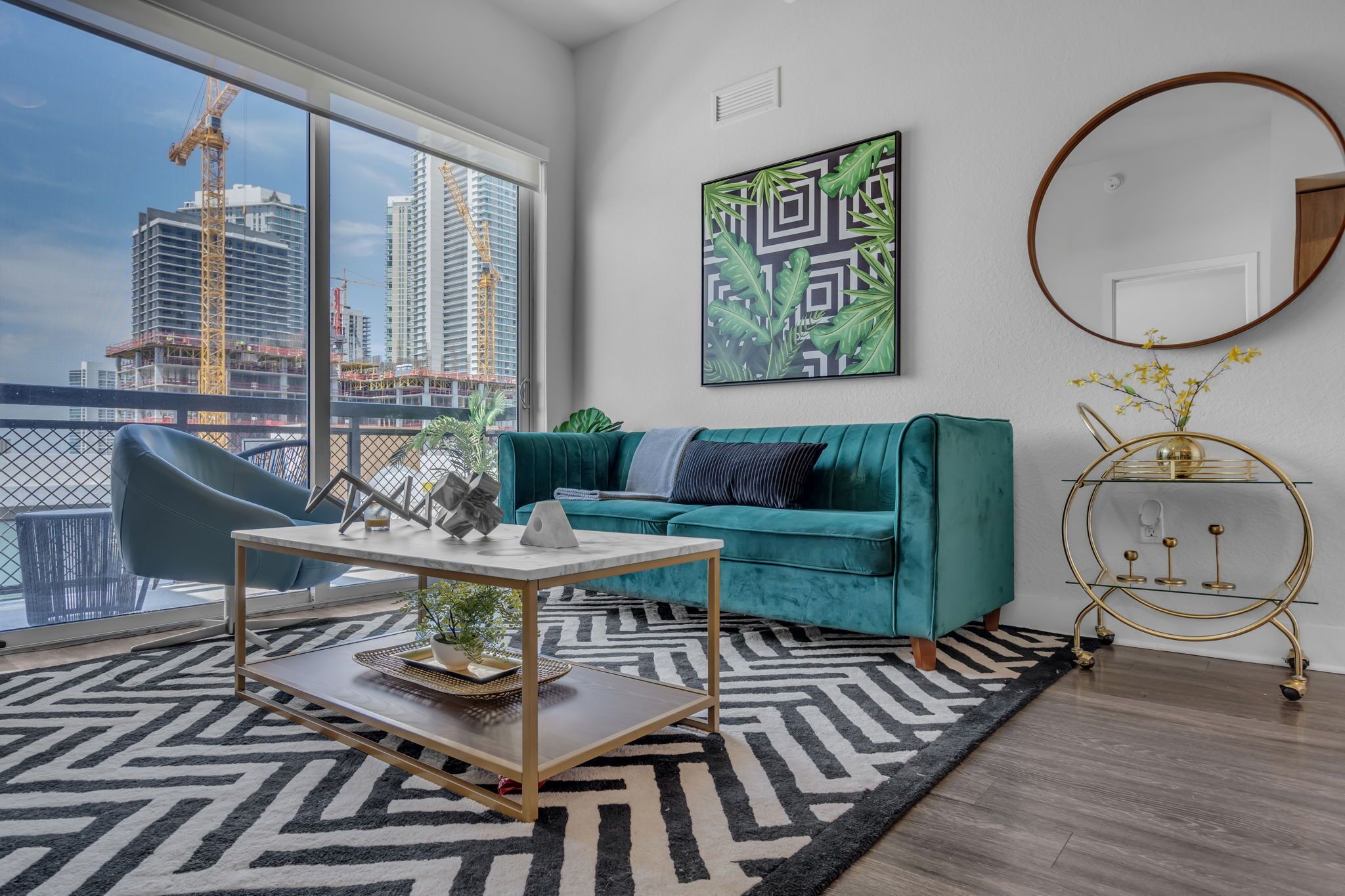 Un salon moderne avec un canapé de couleur turquoise, une table basse, une grande fenêtre donnant sur des immeubles, un tableau abstrait encadré et une table d’appoint dorée.