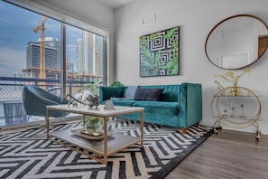 Una sala de estar moderna con un sofá color turquesa, una mesa de centro, un ventanal con vista a edificios, un cuadro abstracto enmarco y una mesita auxiliar dorada.