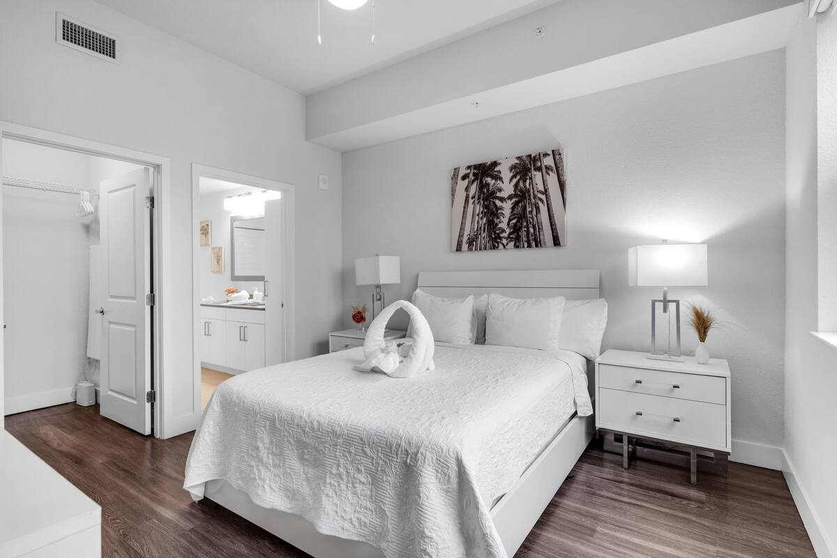 A modern bedroom with a white bed, a nightstand with a lamp, a wall-mounted artwork, and a door leading to another room.