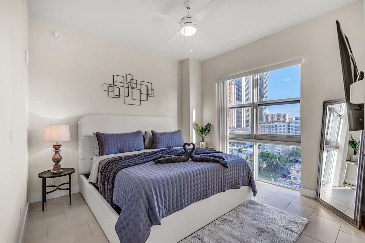 A modern bedroom with a large bed, a side table with a lamp, a decorative wall piece, and a large window with a city view.