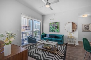 A modern living room with a teal sofa, a coffee table, a round mirror, and a view of the city through large windows. - Blu Apartments (Miami)