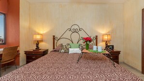 Habitación de hotel con una colcha estampada, un cabecero decorativo, dos mesitas de noche con lámparas, una silla de mimbre y una pequeña mesa con un florero y dos tazas.