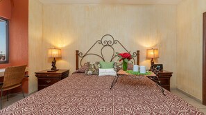 Ein Hotelzimmer mit einem gemusterten Bettbezug, einem dekorativen Kopfteil, zwei Nachttischen mit Lampen, einem Korbstuhl und einem kleinen Tisch mit einer Vase und zwei Tassen.