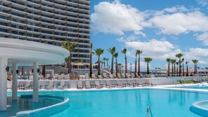 Un espace piscine aménagé dans un complexe hôtelier, avec des chaises longues et des palmiers, entouré d’un grand hôtel.