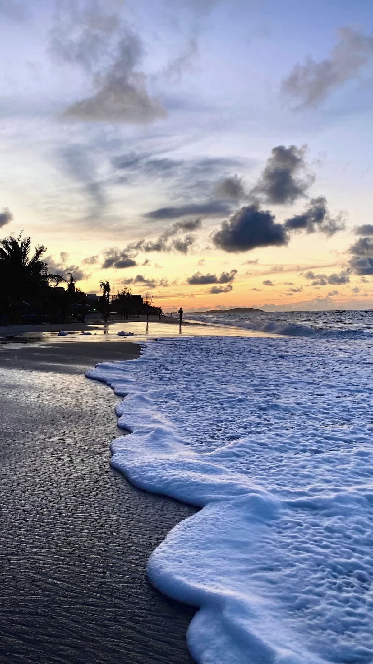 Een strand met golven, een zonsondergang en palmbomen.
