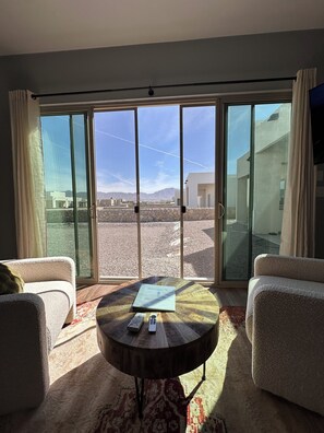 Una habitación con un gran ventanal corredero que da a un balcón, dos sillones, una mesa redonda con un libro y controles remotos, y una alfombra estampada.