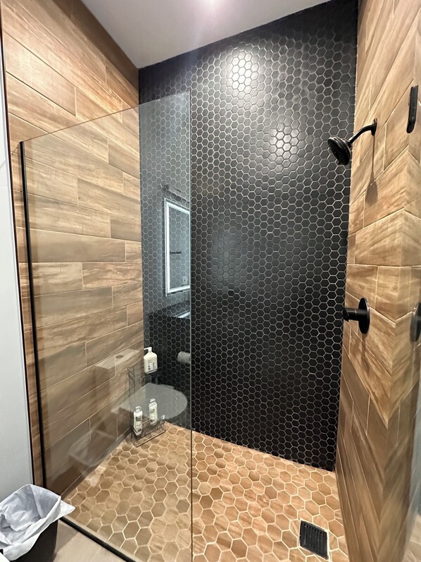 A modern bathroom with a glass shower enclosure, a wooden vanity, and a black hexagonal tile wall.