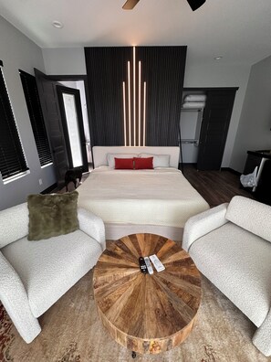 A modern bedroom with a large bed, two white sofas, a round wooden coffee table, and a ceiling light fixture.