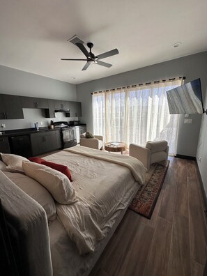 A modern bedroom with a large bed, a sitting area with a coffee table, a kitchenette, and a ceiling fan. - The Desert Light Casita (El Paso)