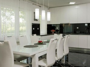 A modern kitchen with white cabinetry, a dining table with chairs, pendant lights, and a view of the outdoors through large windows. - Villa Ante With Outdoor Pool (Imotski)
