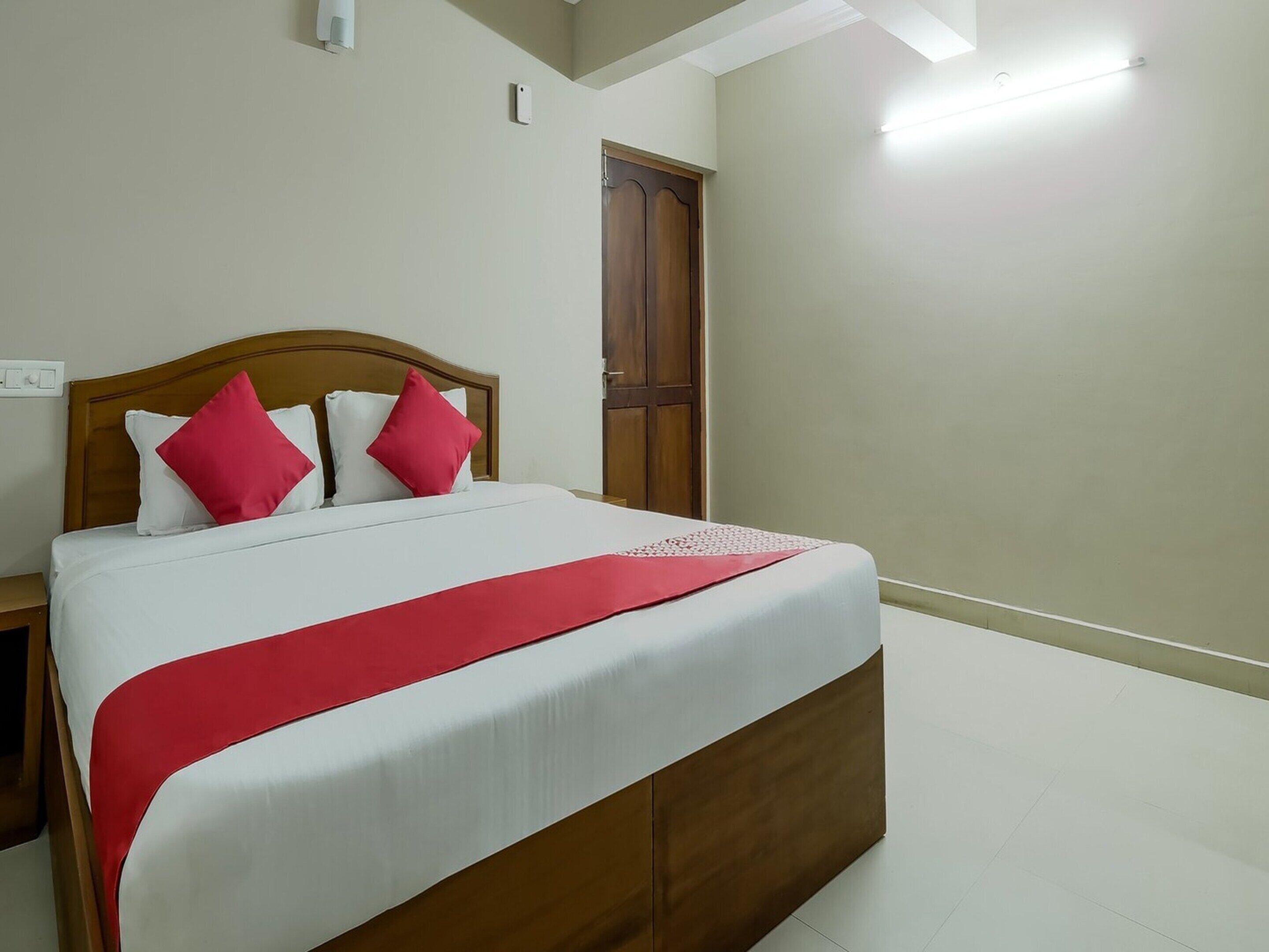 A neatly made bed with a wooden headboard, white bedding, and red accents, in a room with a wooden door and a light switch on the wall.