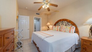 A bedroom with a bed, wooden headboard, bedside lamps, a ceiling fan, a wooden dresser, and a door leading to another room. - Beautiful Beachfront Retreat With Panoramic Ocean Views (Backus)
