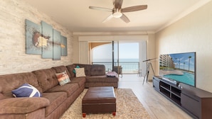 A living room with a brown sofa, a flat-screen TV, a ceiling fan, and a balcony with a pool view. - Stunning Oceanfront Escape With Sweeping Sea Views (Puerto Peñasco)