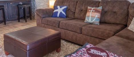 A brown leather sofa with two decorative pillows, a matching ottoman, and a patterned throw on a furry rug.