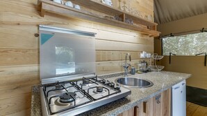 A kitchen with a gas stove, a sink, wooden shelves with kitchenware, and a window with a view. - Boavista Glamping Tent with Mountain View, Private Pool and Wi-Fi (Arco de Baúlhe)
