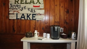 Una pared de madera con un letrero que dice 'RELAX YOU'RE AT THE LAKE' y una mesa con una cafetera, tetera, tazas y una cesta.