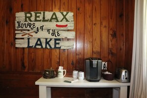 A wooden wall with a sign that reads 'RELAX YOU'RE AT THE LAKE' and a table with a coffee maker, teapot, cups, and a basket.