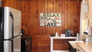 Una cocina con paredes de madera, ventilador de techo, refrigerador, cafetera y un letrero que dice 'RELAX Your time at the LAKE'.
