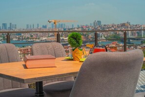Una mesa con un libro, una planta pequeña y un bolso amarillo en un balcón con vistas a un paisaje urbano que incluye edificios y un puente.