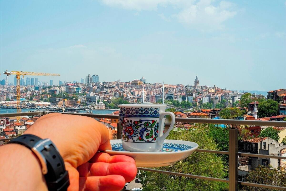 Een hand houdt een beker met een decoratief patroon vast, met uitzicht op een stadsgezicht met gebouwen en een waterpartij.
