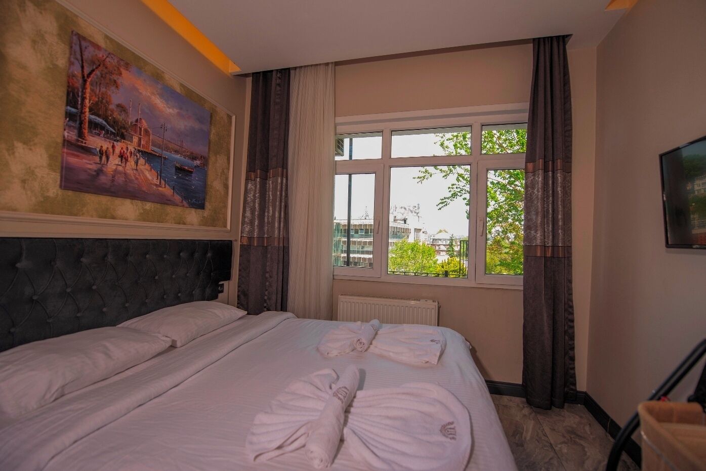 Habitación de hotel con una cama grande, un cuadro en la pared, una ventana con cortinas y un radiador.