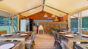 A tented dining area with wooden tables and benches, a kitchen in the background, and large windows.