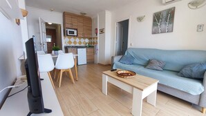 Ein Wohnzimmer mit einer Couch, einem Couchtisch und einem Fernseher. Es gibt eine Küchenzeile mit Schränken und einer Mikrowelle.