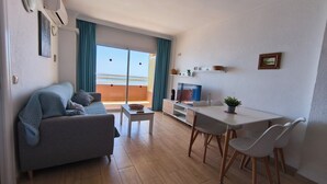 Ein modernes Wohnzimmer mit einer Couch, einem Esstisch mit Stühlen, einem Fernseher und einem Balkon mit Vorhängen.