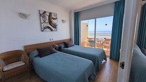 Ein Hotelzimmer mit zwei Betten, einem Korbstuhl, einem Bild an der Wand und einem Balkon mit Meerblick.