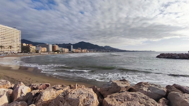 Ein Strand mit felsigem Vordergrund, einem Sandufer und einer Stadtlandschaft mit Gebäuden und Bergen im Hintergrund.