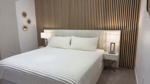 Un lit bien fait, recouvert d’une couvre-lit blanche et agrémenté d’oreillers, avec des tables de chevet et des lampes de chaque côté, le tout adossé à un mur orné de lattes de bois verticales.