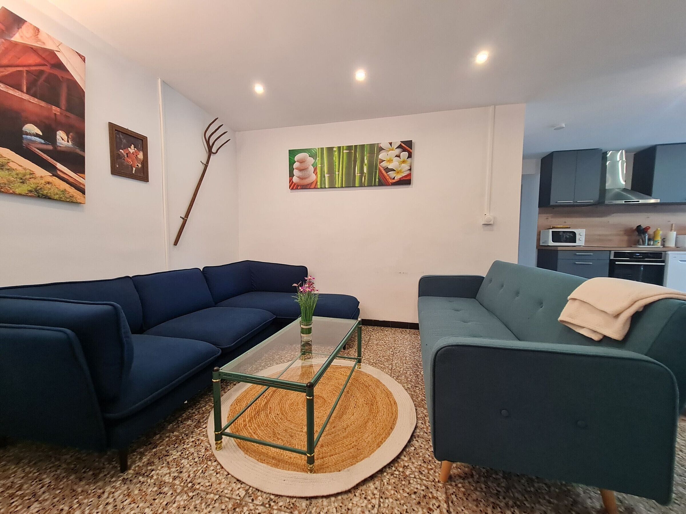 A living room with a blue sofa, a green sofa, a glass coffee table, and a kitchen area with cabinets and appliances.