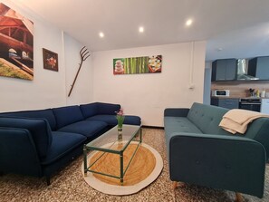 Ein Wohnzimmer mit blauem Sofa, grünem Sofa, einem Glastisch und einer Küchenzeile mit Schränken und Geräten.