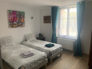 Une chambre avec deux lits simples, une petite table avec une lampe, une horloge murale, une fenĂȘtre avec des rideaux et un tableau au mur.