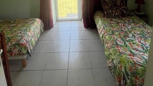 Una habitación con dos camas, ventilador de techo, ventana con cortinas y suelo de baldosas.