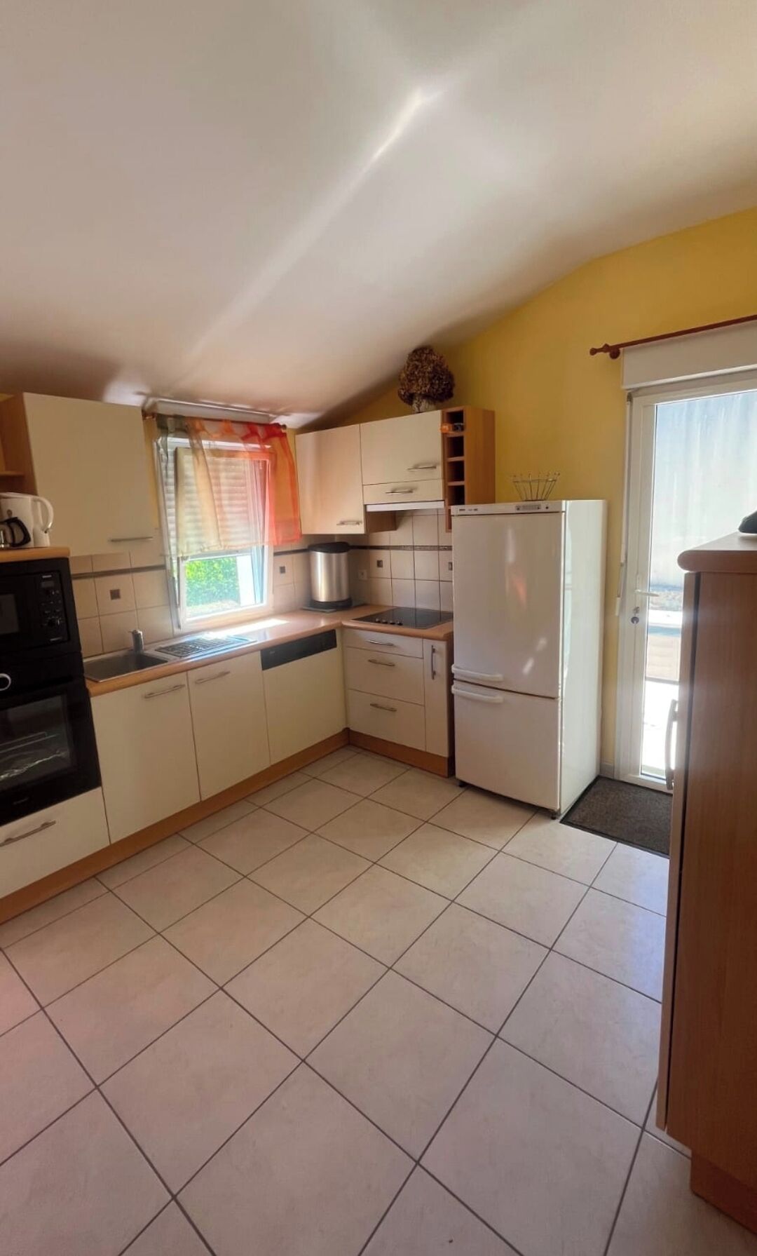 Une cuisine compacte avec des meubles blancs, un four encastré, un réfrigérateur et une fenêtre avec un rideau.