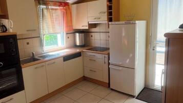 Une cuisine compacte avec des meubles blancs, un four encastré, un réfrigérateur et une fenêtre avec un rideau.