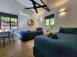 A bedroom with two beds, a desk, a chair, a ceiling fan, and a window with a view of greenery.