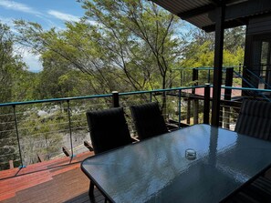 A terrace with a glass table and chairs, a metal railing, and a view of trees and a distant landscape.