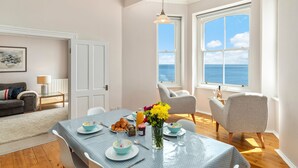 Un coin repas avec une table mise pour le petit-déjeuner, un vase rempli de fleurs et une vue sur la mer par de grandes fenêtres.