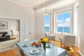 Ein Essbereich mit einem für das Frühstück gedeckten Tisch, einer Blumenvase und Meerblick durch große Fenster.