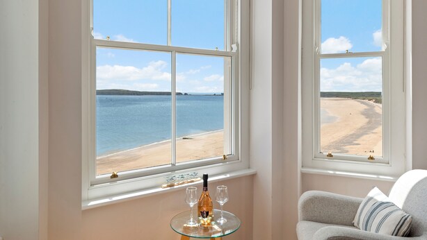 Une pièce avec vue sur la plage, grâce à de grandes fenêtres, une table en verre avec une bouteille et des verres, et un canapé gris agrémenté d’un coussin à rayures.