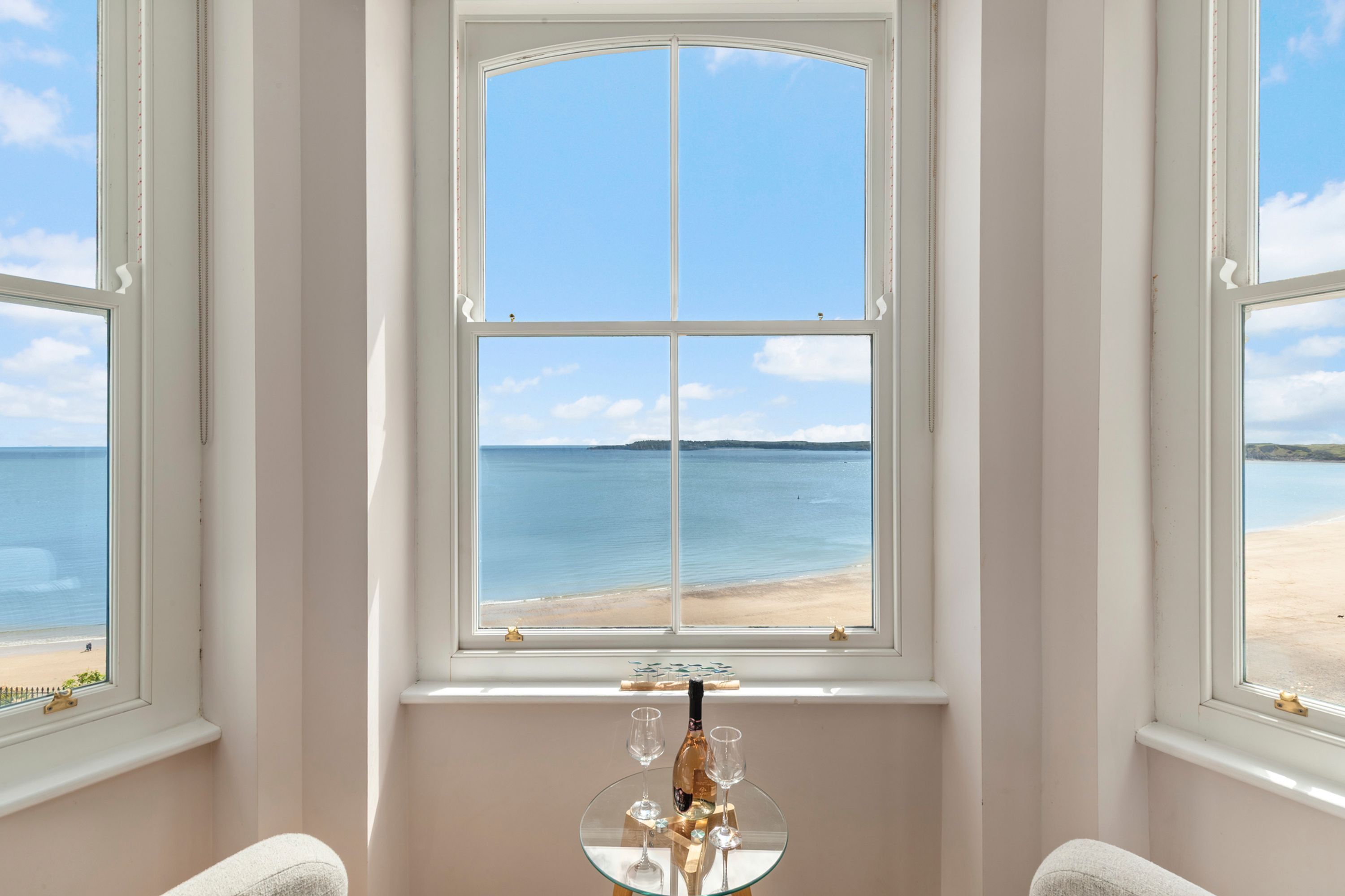 A room with a large window overlooking a beach, a small table with a bottle and glasses, and a chair.
