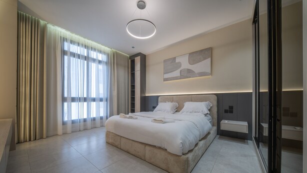 A modern bedroom with a large bed, a bedside table, a built-in shelf, and a large window with curtains. - A comfortable 2 bedroom apartment in Al-Hamra (Jeddah)
