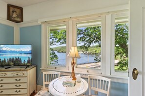 Una habitación con una televisión que muestra un lago de montaña, una mesa con una lámpara y pañuelos, y vistas a un lago y árboles a través de ventanas amplias.